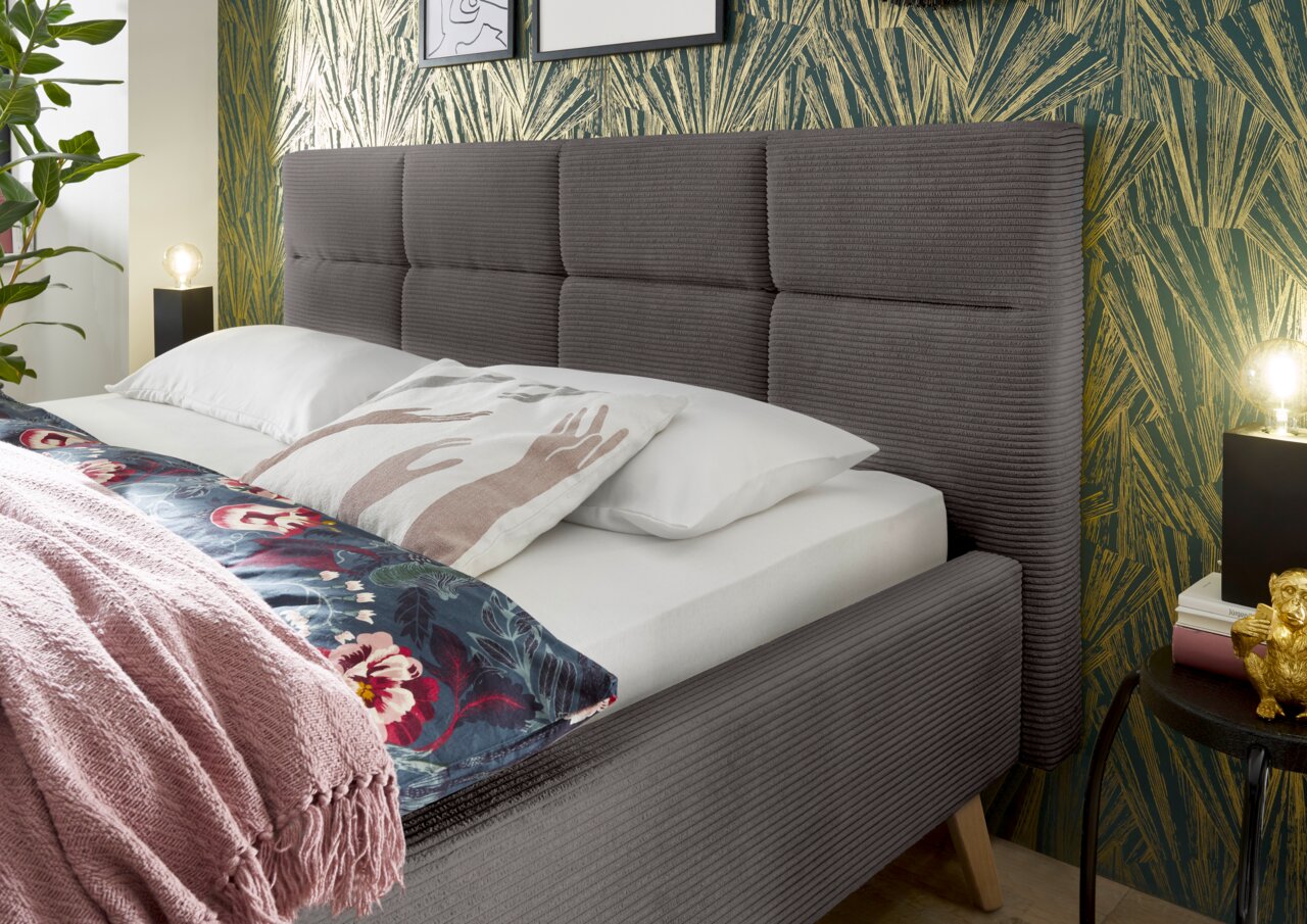 Zijaanzicht van een modern gestoffeerd bed met een grijs hoofdeinde, versierd met kussens en een kleurrijke deken, tegen een stijlvol behang.