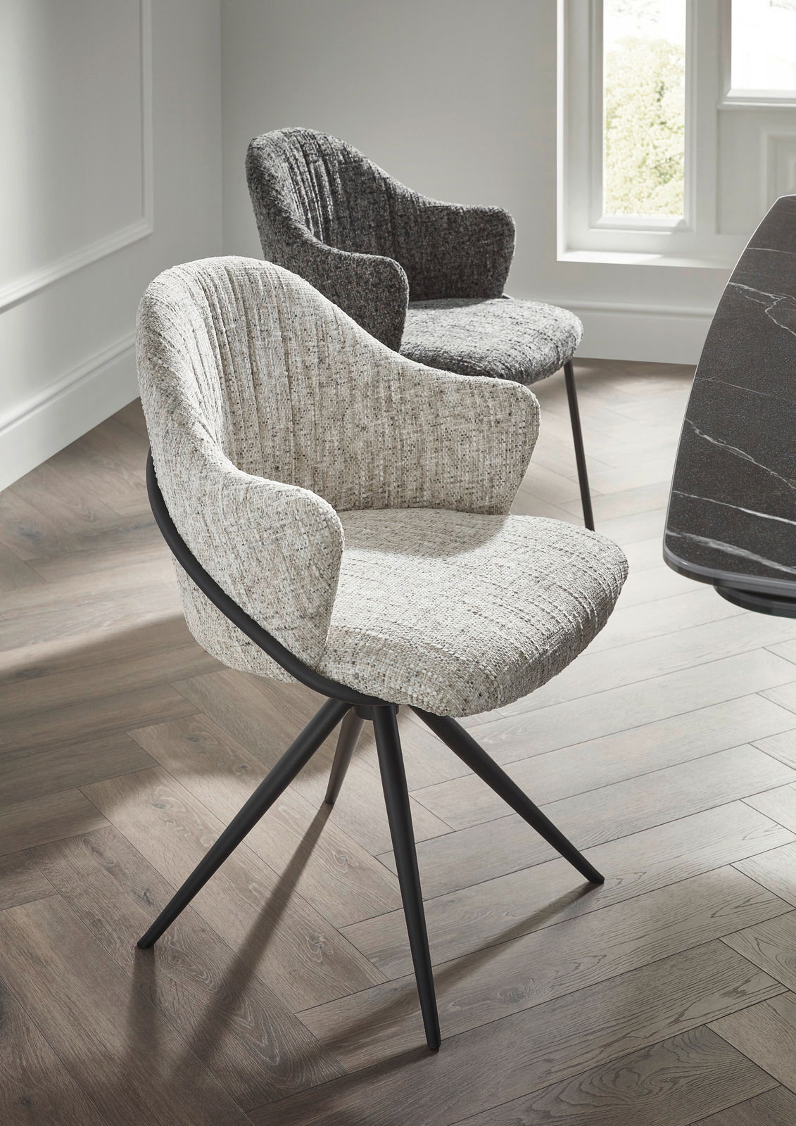 Zwei moderne Armlehnstühle mit Stoffbezug in hellen und dunklen Grautönen, auf einem Holzfußboden in einem hellen Raum, aus seitlicher Perspektive fotografiert.