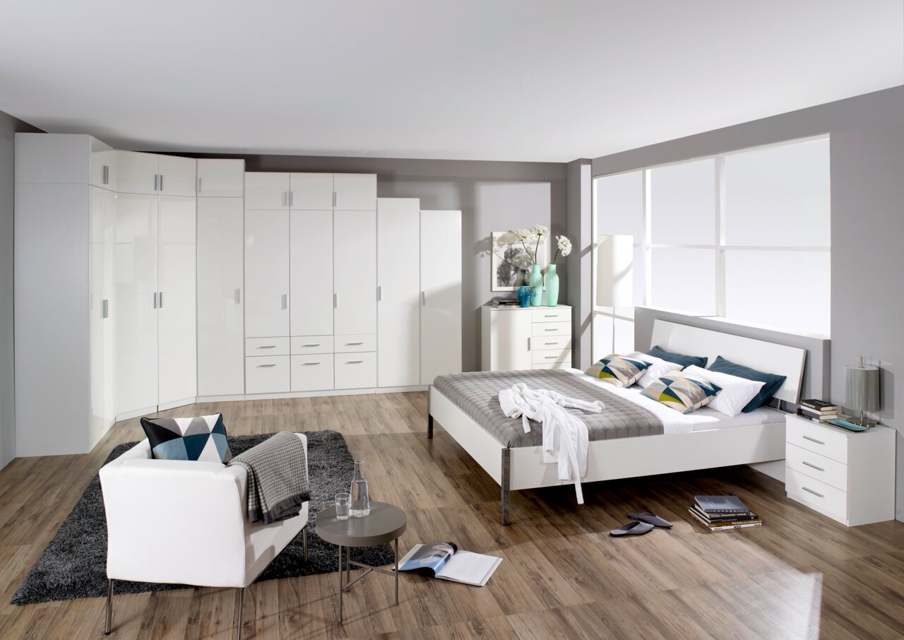 Moderne Schlafzimmermöbel in Weiß, bestehend aus einem großen Kleiderschrank mit Drehtüren, einem Bett mit grauer Bettwäsche und Nachttischen, aus der Perspektive einer Raumansicht.