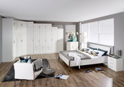 Moderne Schlafzimmermöbel in Weiß, bestehend aus einem großen Kleiderschrank mit Drehtüren, einem Bett mit grauer Bettwäsche und Nachttischen, aus der Perspektive einer Raumansicht.