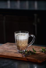 Eleganter Barista Kaffeebecher mit Henkel, gefüllt mit einem Schichtgetränk aus Kaffee und Milchschaum, auf einem Holzbrett mit dekorativen Kräutern, seitliche Perspektive.