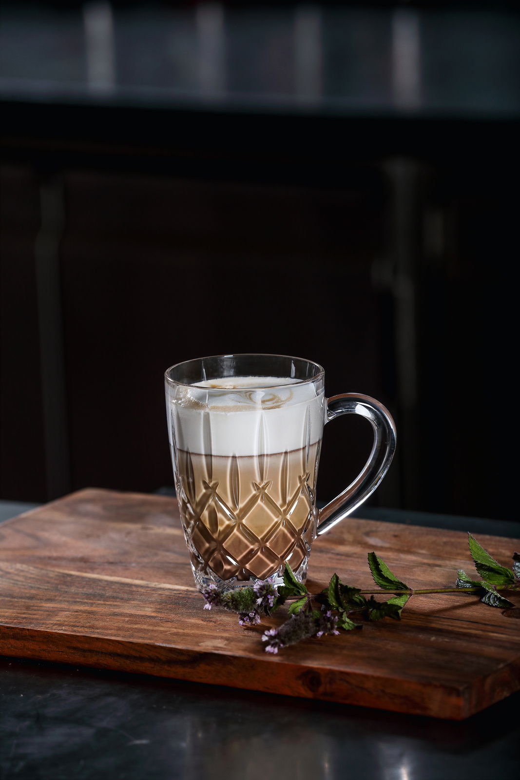 Eleganter Barista Kaffeebecher mit Henkel, gefüllt mit einem Schichtgetränk aus Kaffee und Milchschaum, auf einem Holzbrett mit dekorativen Kräutern, seitliche Perspektive.