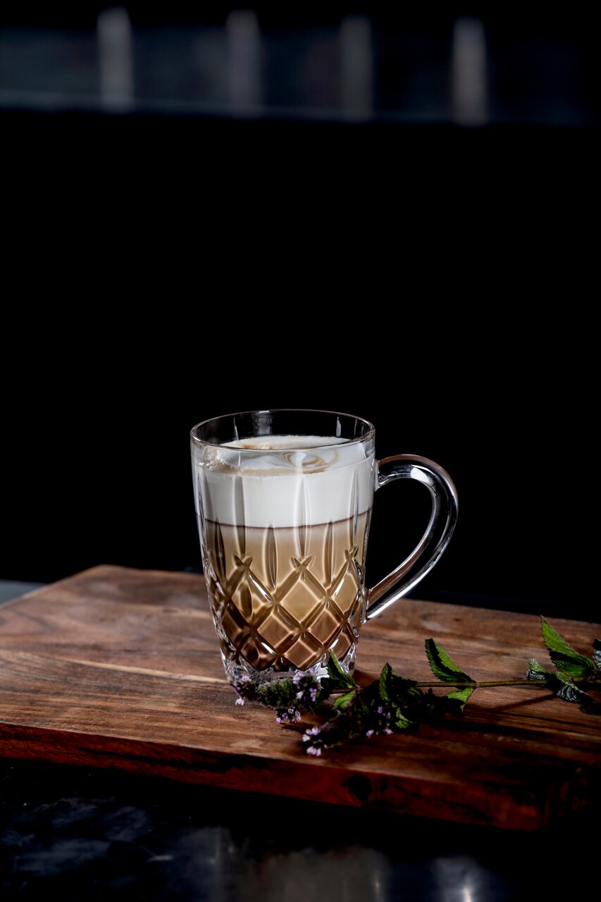 Eleganter Barista Kaffeebecher mit Henkel, gefüllt mit einem Schichtgetränk aus Kaffee und Milchschaum, auf einem Holzbrett mit dekorativen Kräutern, seitliche Perspektive.