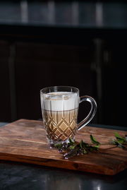 Eleganter Barista Kaffeebecher mit Henkel, gefüllt mit einem Schichtgetränk aus Kaffee und Milchschaum, auf einem Holzbrett mit dekorativen Kräutern, seitliche Perspektive.