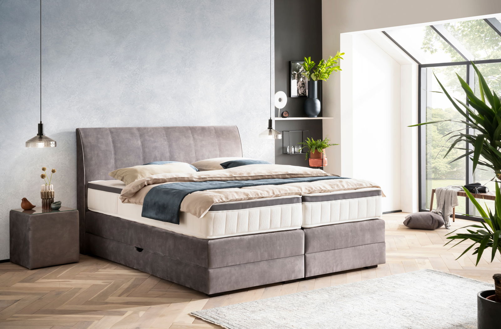 Boxspringbett in einem modernen Schlafzimmer mit grauem Kopfteil, seitlicher Perspektive, umgeben von Pflanzen und natürlichem Licht.