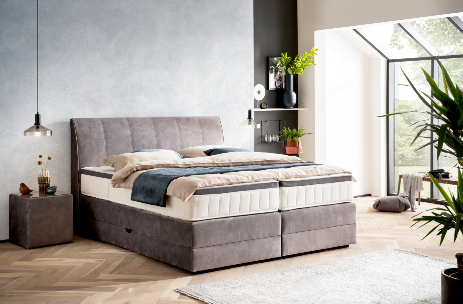 Boxspringbett in einem modernen Schlafzimmer mit grauem Kopfteil, seitlicher Perspektive, umgeben von Pflanzen und natürlichem Licht.