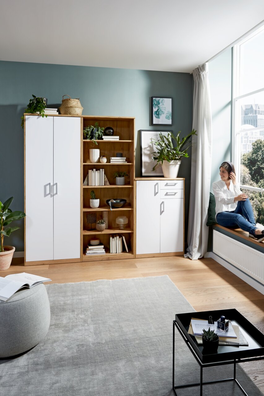 Wohnzimmer mit einem weißen Kleiderschrank, einem offenen Holzregal und einer weißen Kommode, seitliche Perspektive