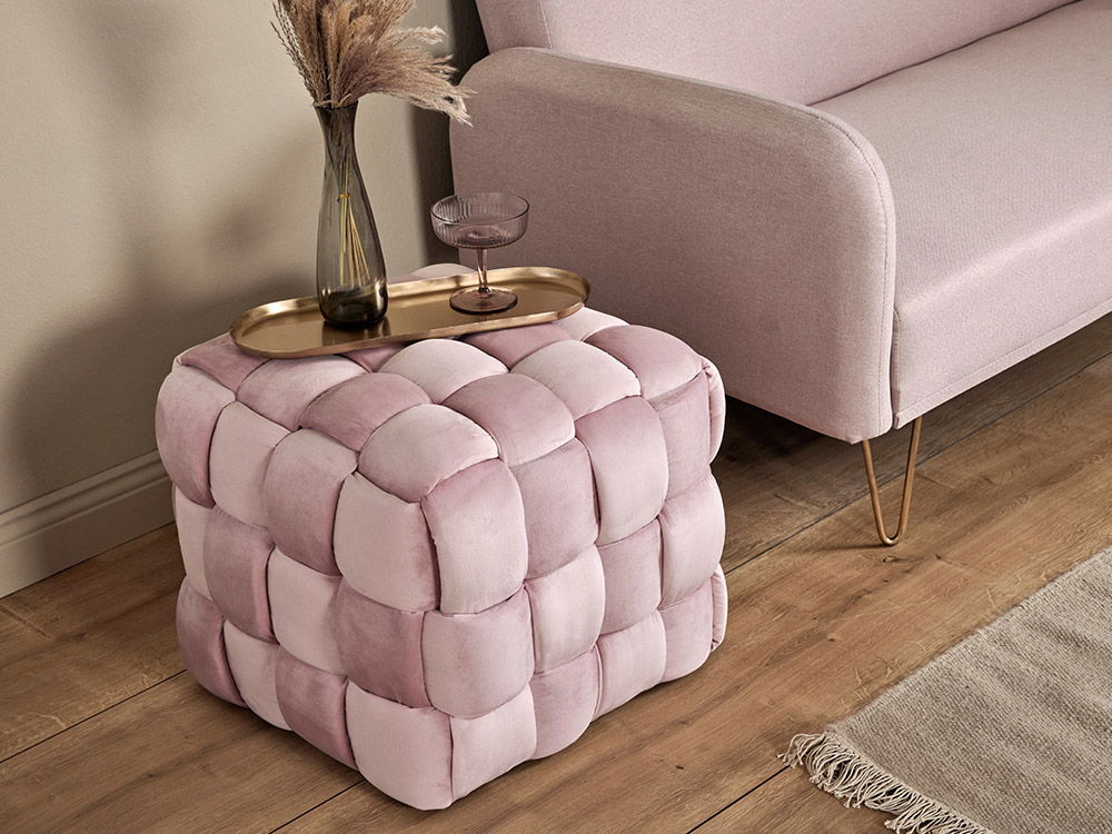 Ein rosafarbener, gewebter Hocker neben einem Sofa mit einem Messingtablett, auf dem eine Vase und ein Glas stehen.