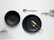 Draufsicht auf schwarzes, strukturiertes Geschirrset mit großer Schale und zwei Tellern, kombiniert mit goldenem Besteck auf hellem Hintergrund