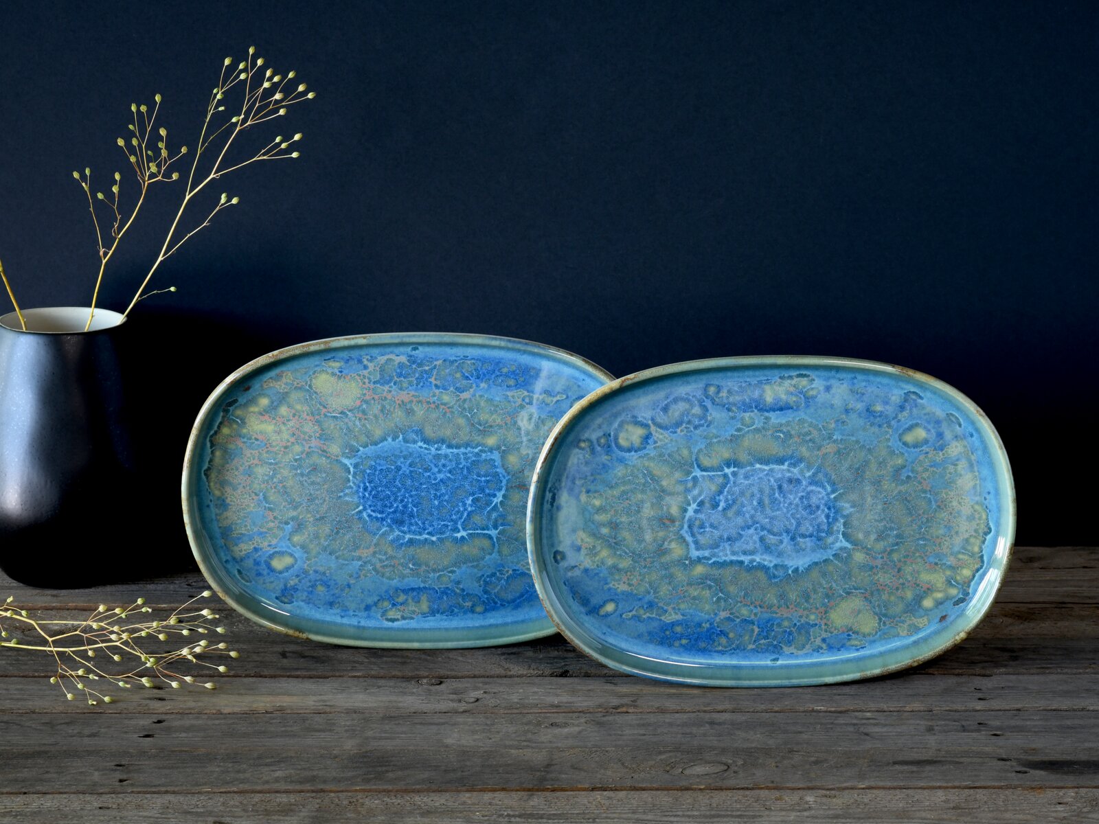 Zwei ovale Keramik-Servierplatten in Blau-Grün mit reaktiver Glasur und organischem Muster, frontal in leichter Schrägansicht auf einem rustikalen Holztisch vor dunklem Hintergrund, links eine schwarze Vase mit zarten Zweigen.