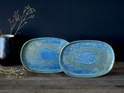 Zwei ovale Keramik-Servierplatten in Blau-Grün mit reaktiver Glasur und organischem Muster, frontal in leichter Schrägansicht auf einem rustikalen Holztisch vor dunklem Hintergrund, links eine schwarze Vase mit zarten Zweigen.