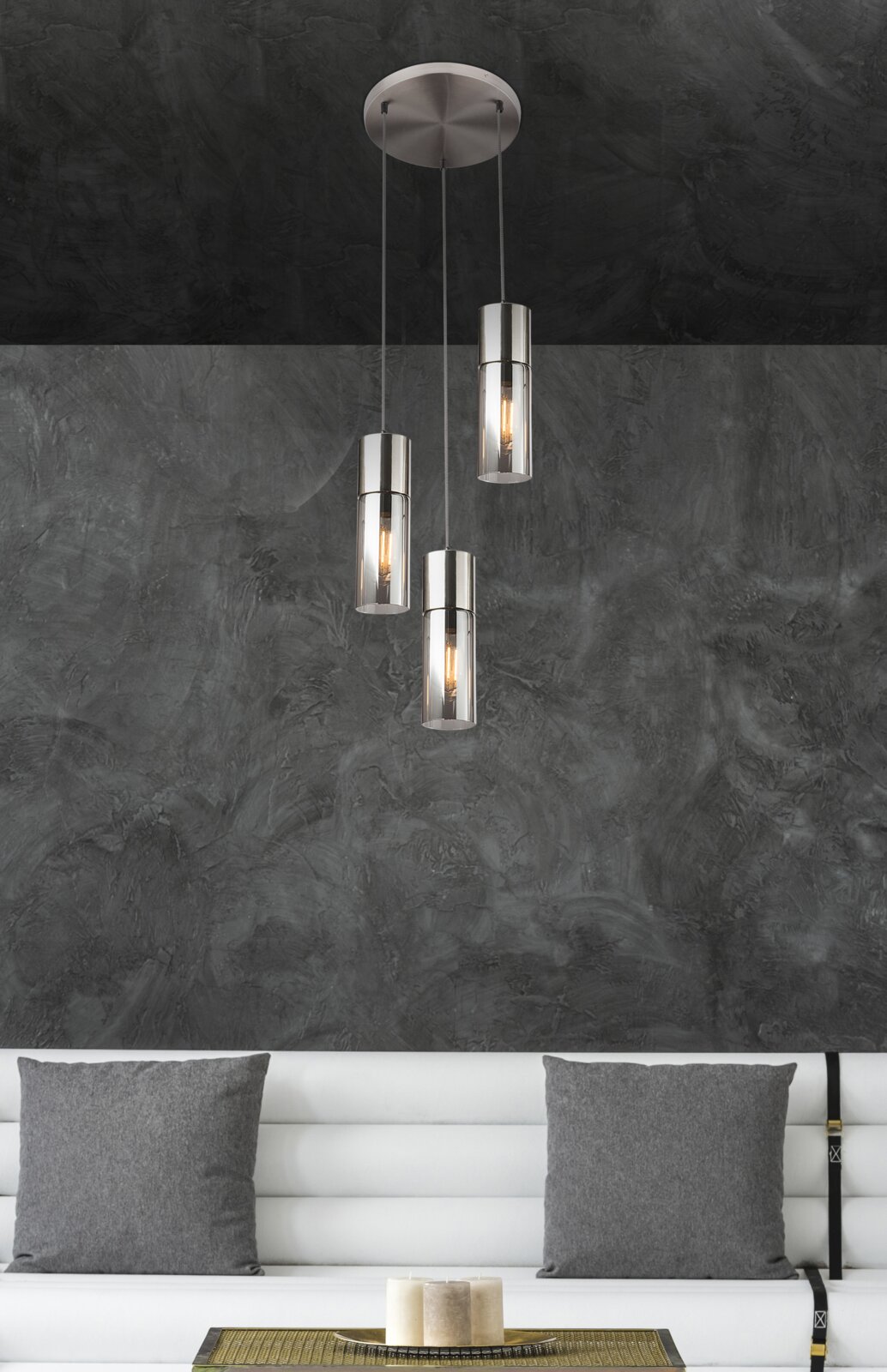 Moderne Hängeleuchte mit drei zylindrischen Lampenschirmen aus Metall und Glas, von der Decke hängend, in einem stilvollen Wohnzimmer mit grauer Wand und weißem Sofa. Perspektive von vorne.