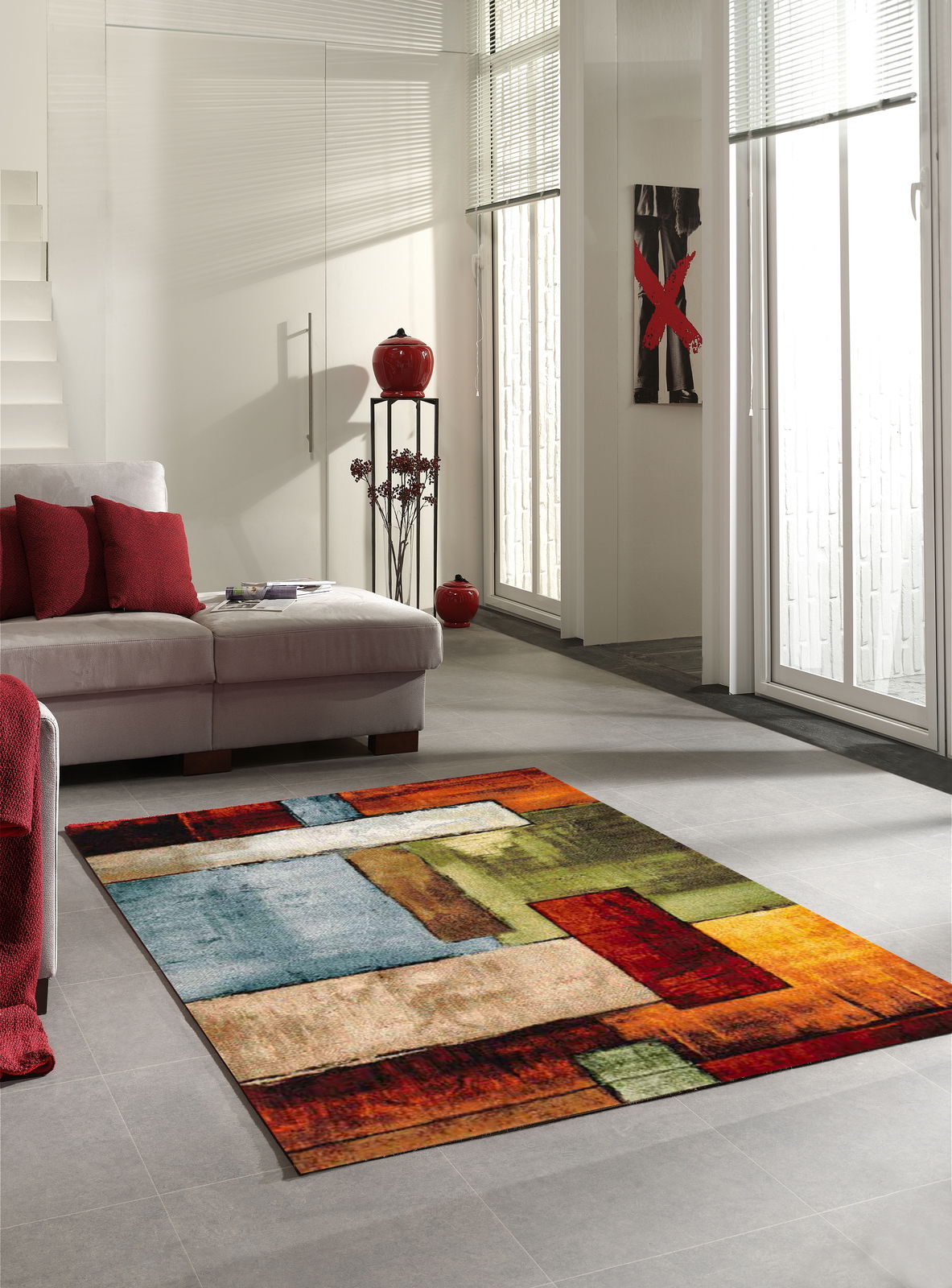 Merinos Webteppich DIAMOND Moderner Teppich mit geometrischen Mustern in lebendigen Farben wie Rot, Grün und Blau, auf einem hellen Wohnzimmerboden vor einem Sofa mit roten Kissen, aus einer seitlichen Perspektive fotografiert.