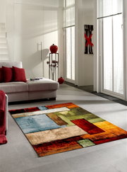 Moderner Teppich mit geometrischen Mustern in lebendigen Farben wie Rot, Grün und Blau, auf einem hellen Wohnzimmerboden vor einem Sofa mit roten Kissen, aus einer seitlichen Perspektive fotografiert.