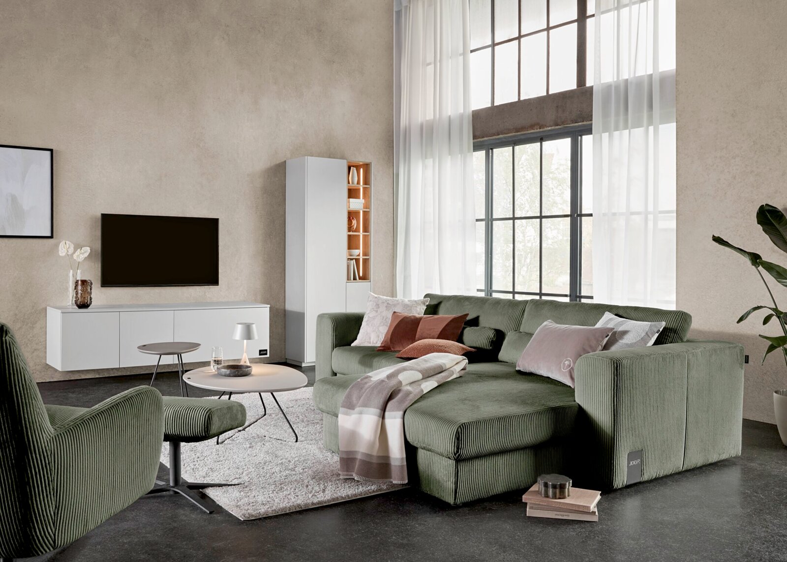 Groene ribfluwelen hoekbank met chaise longue, sierkussens en plaid in een lichte moderne woonkamer, schuin van voren gezien.
