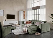 Groene ribfluwelen hoekbank met chaise longue, sierkussens en plaid in een lichte moderne woonkamer, schuin van voren gezien.