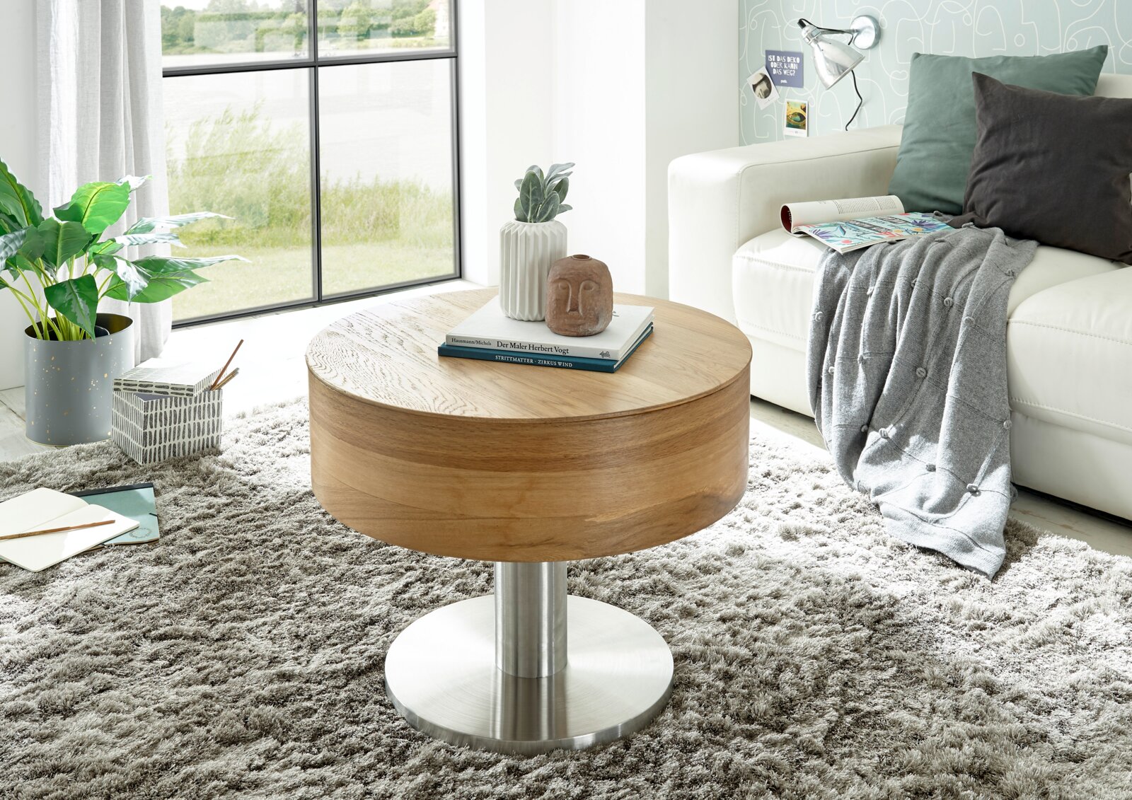 Moderne ronde houten salontafel met metalen onderstel, versierd met boeken en een vaas, in een gezellige woonkamer met tapijt en bank, zijaanzicht.