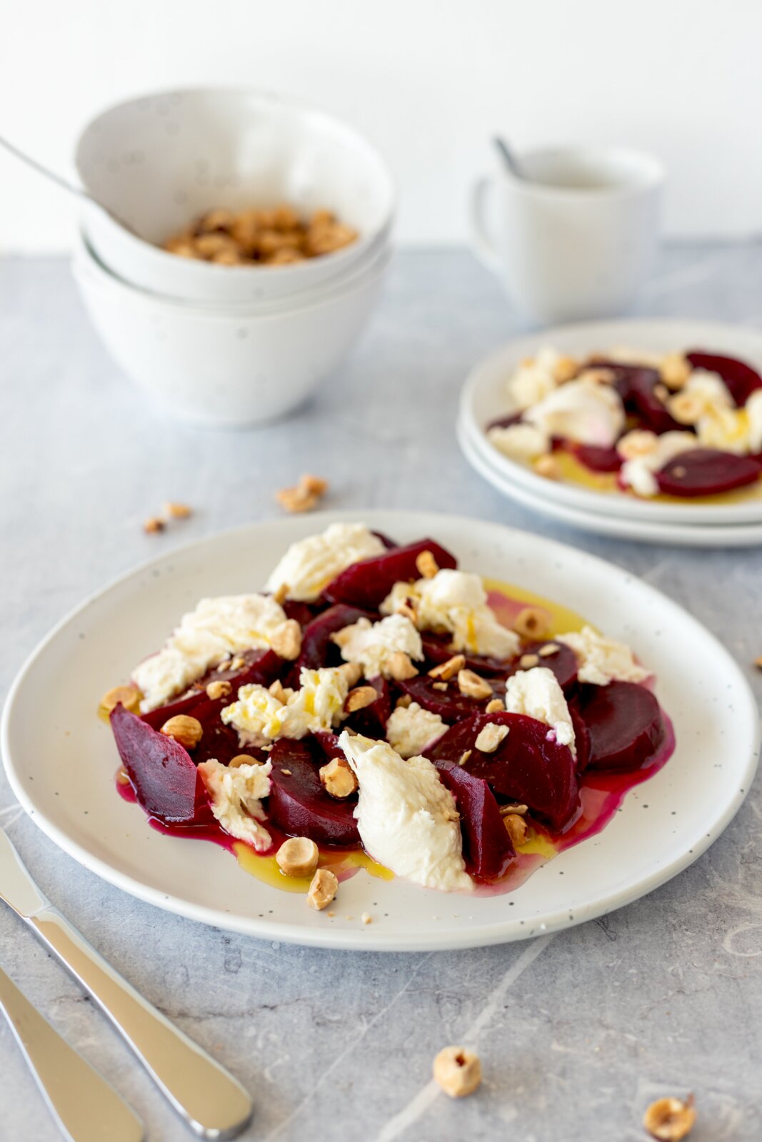 Nahaufnahme in leicht schräger Draufsicht: Rote-Bete-Salat mit cremigem Käse, gehackten Nüssen und Olivenöl auf weißem Teller; im Hintergrund Schale und Tasse