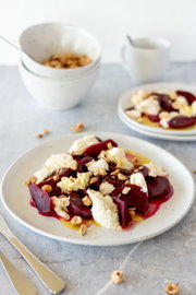 Nahaufnahme in leicht schräger Draufsicht: Rote-Bete-Salat mit cremigem Käse, gehackten Nüssen und Olivenöl auf weißem Teller; im Hintergrund Schale und Tasse