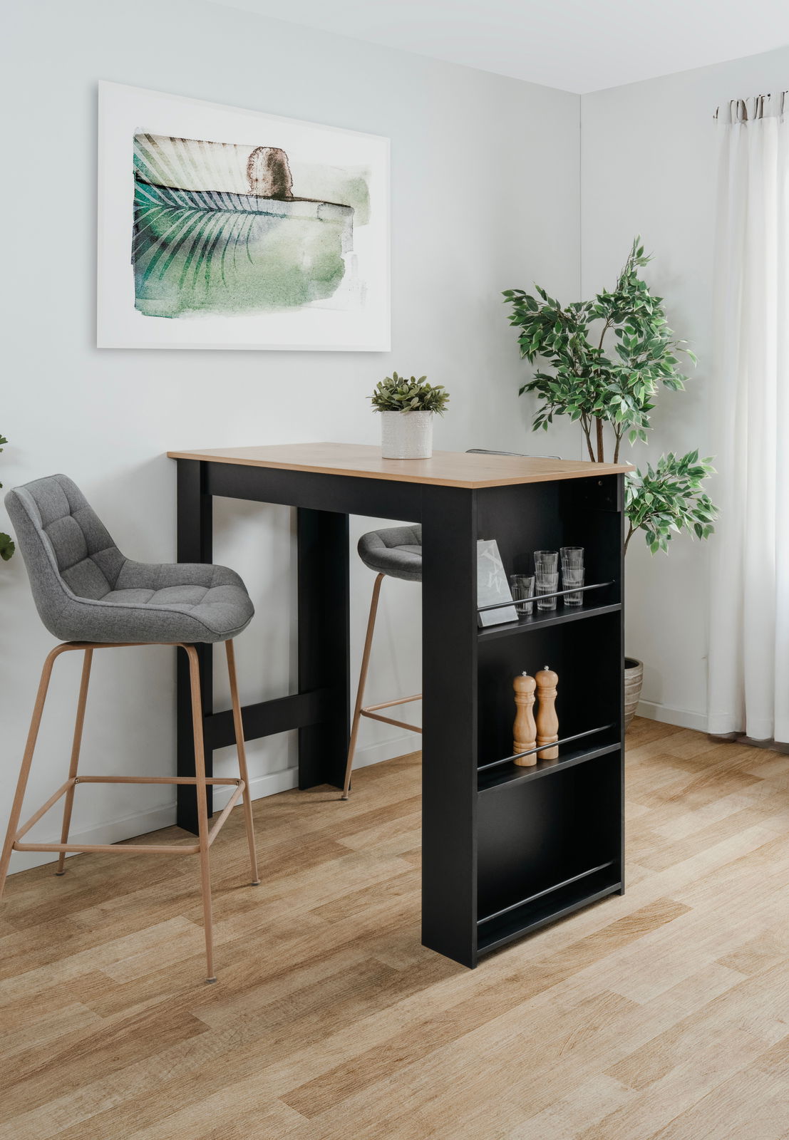 Moderner Bartisch mit schwarzem Gestell und Holzplatte, umgeben von zwei grauen Barhockern, in einem hellen Raum mit Pflanzen und Wandkunst. Perspektive von schräg vorne.
