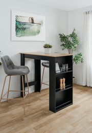 Moderner Bartisch mit schwarzem Gestell und Holzplatte, umgeben von zwei grauen Barhockern, in einem hellen Raum mit Pflanzen und Wandkunst. Perspektive von schräg vorne.