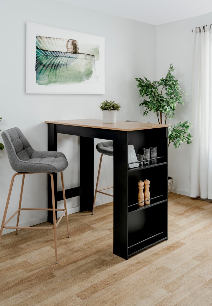 Moderner Bartisch mit schwarzem Gestell und Holzplatte, umgeben von zwei grauen Barhockern, in einem hellen Raum mit Pflanzen und Wandkunst. Perspektive von schräg vorne.