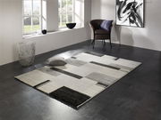 Moderner Teppich in Grau- und Schwarztönen mit geometrischem Muster, in einem stilvollen Raum mit Fenster, Sessel und Dekorationen, aus der Perspektive von schräg oben betrachtet.
