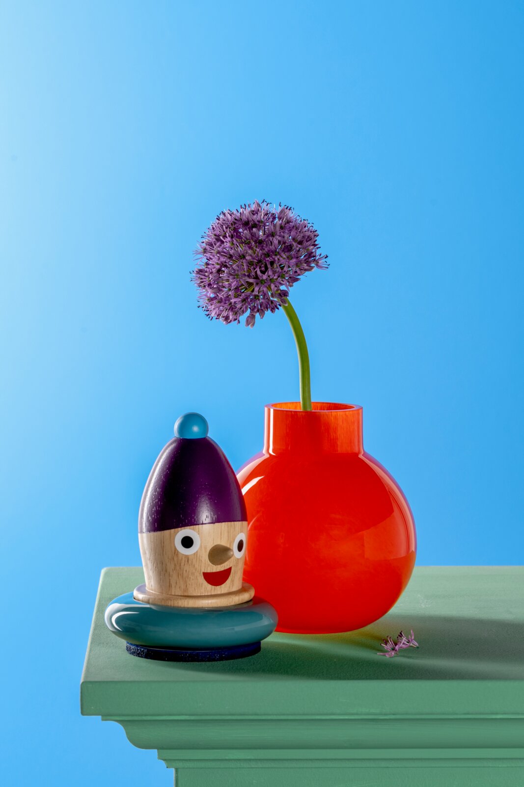 Rote Vase mit rundem Körper und schmalem Hals, gefüllt mit einer lila Blume, auf einem grünen Tisch vor blauem Hintergrund. Perspektive von vorne.