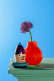 Rote Vase mit rundem Körper und schmalem Hals, gefüllt mit einer lila Blume, auf einem grünen Tisch vor blauem Hintergrund. Perspektive von vorne.