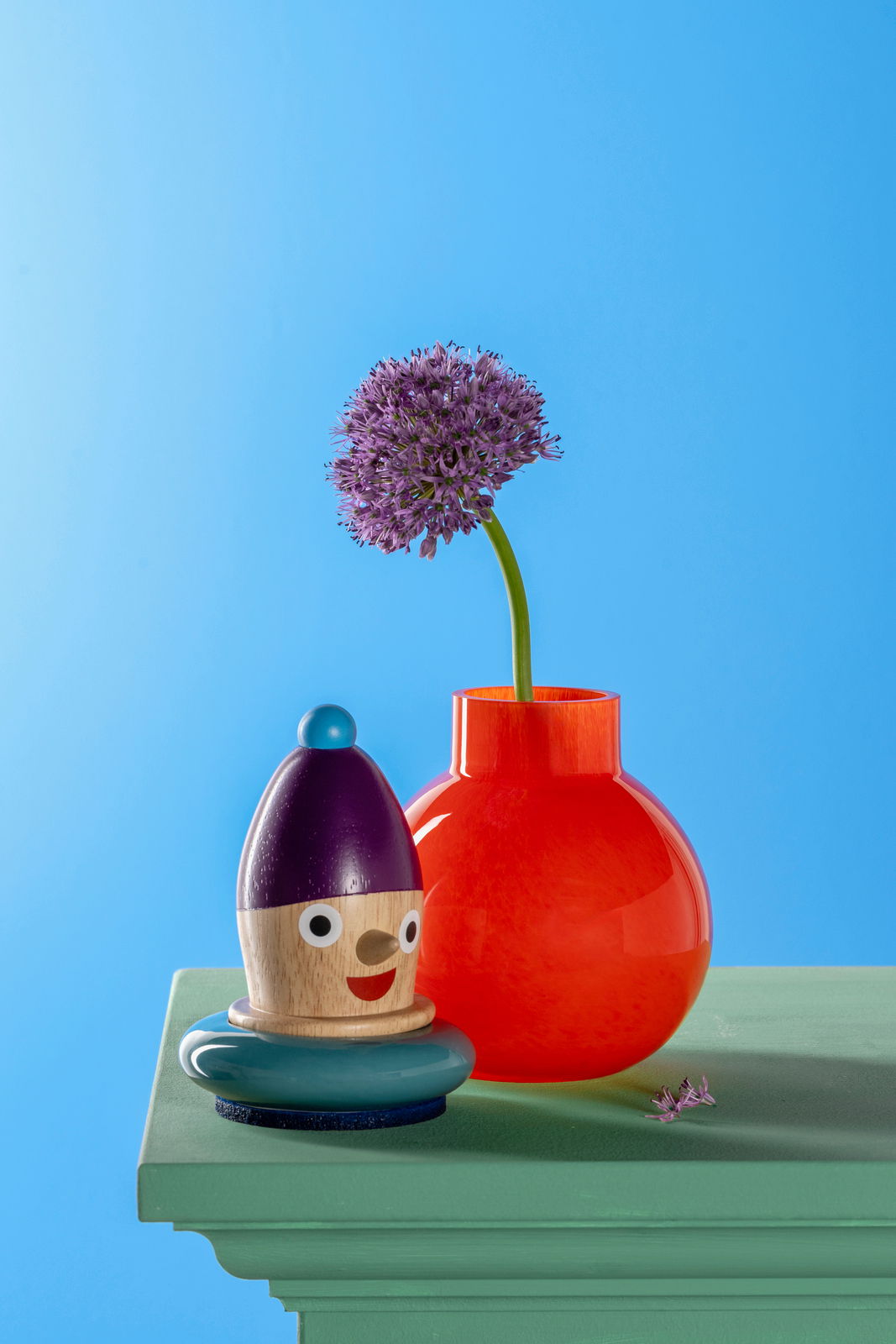 Rote Vase mit rundem Körper und schmalem Hals, gefüllt mit einer lila Blume, auf einem grünen Tisch vor blauem Hintergrund. Perspektive von vorne.