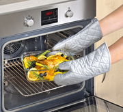 Person mit silbernen Ofenhandschuhen nimmt eine Glasform mit gebratenem Hähnchen, Zitronenscheiben und Kräutern aus einem modernen Backofen. Perspektive: Frontalansicht auf den Ofen.