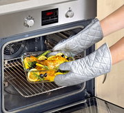 Person mit silbernen Ofenhandschuhen nimmt eine Glasform mit gebratenem Hähnchen, Zitronenscheiben und Kräutern aus einem modernen Backofen. Perspektive: Frontalansicht auf den Ofen.