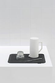 Schwarze Abtropfmatte auf weißer Oberfläche mit einem umgedrehten Glas, einem weißen Krug und einem Messer, aus der Seitenperspektive fotografiert.