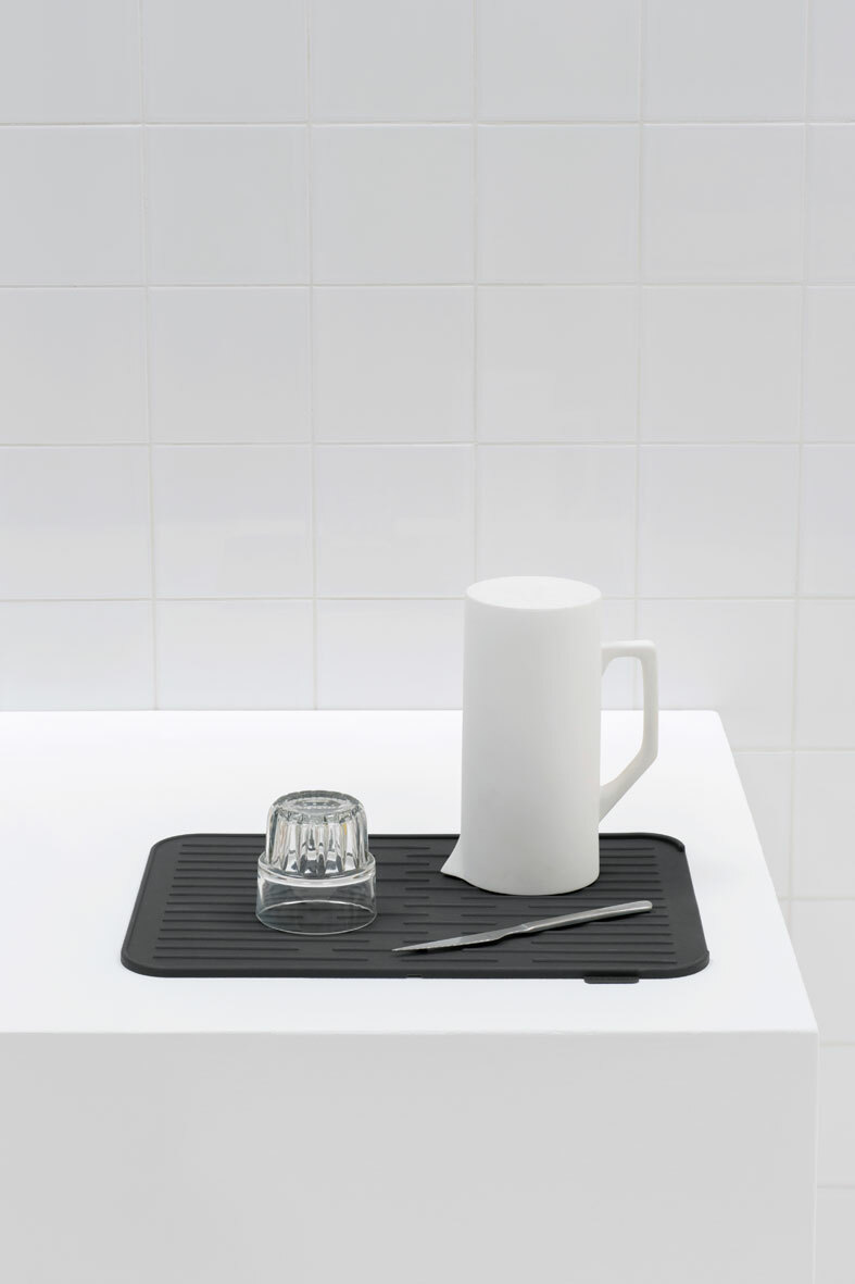 Schwarze Abtropfmatte auf weißer Oberfläche mit einem umgedrehten Glas, einem weißen Krug und einem Messer, aus der Seitenperspektive fotografiert.