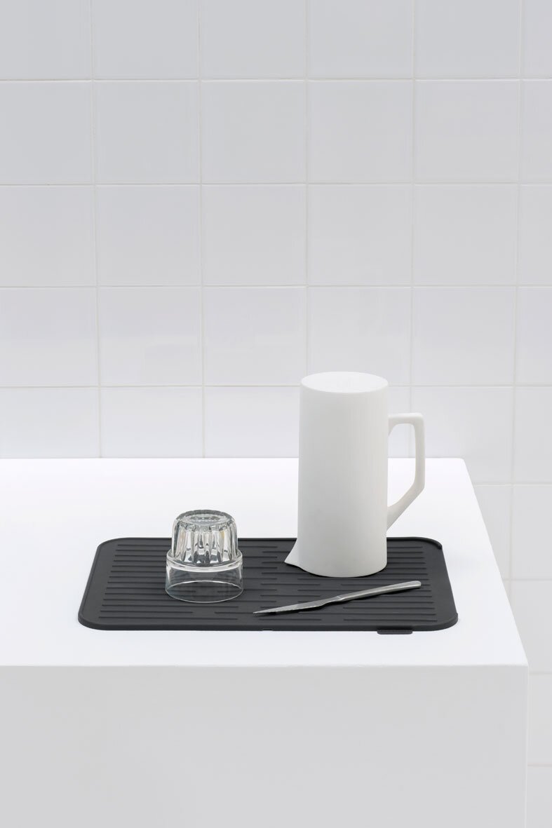 Schwarze Abtropfmatte auf weißer Oberfläche mit einem umgedrehten Glas, einem weißen Krug und einem Messer, aus der Seitenperspektive fotografiert.