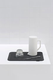 Schwarze Abtropfmatte auf weißer Oberfläche mit einem umgedrehten Glas, einem weißen Krug und einem Messer, aus der Seitenperspektive fotografiert.