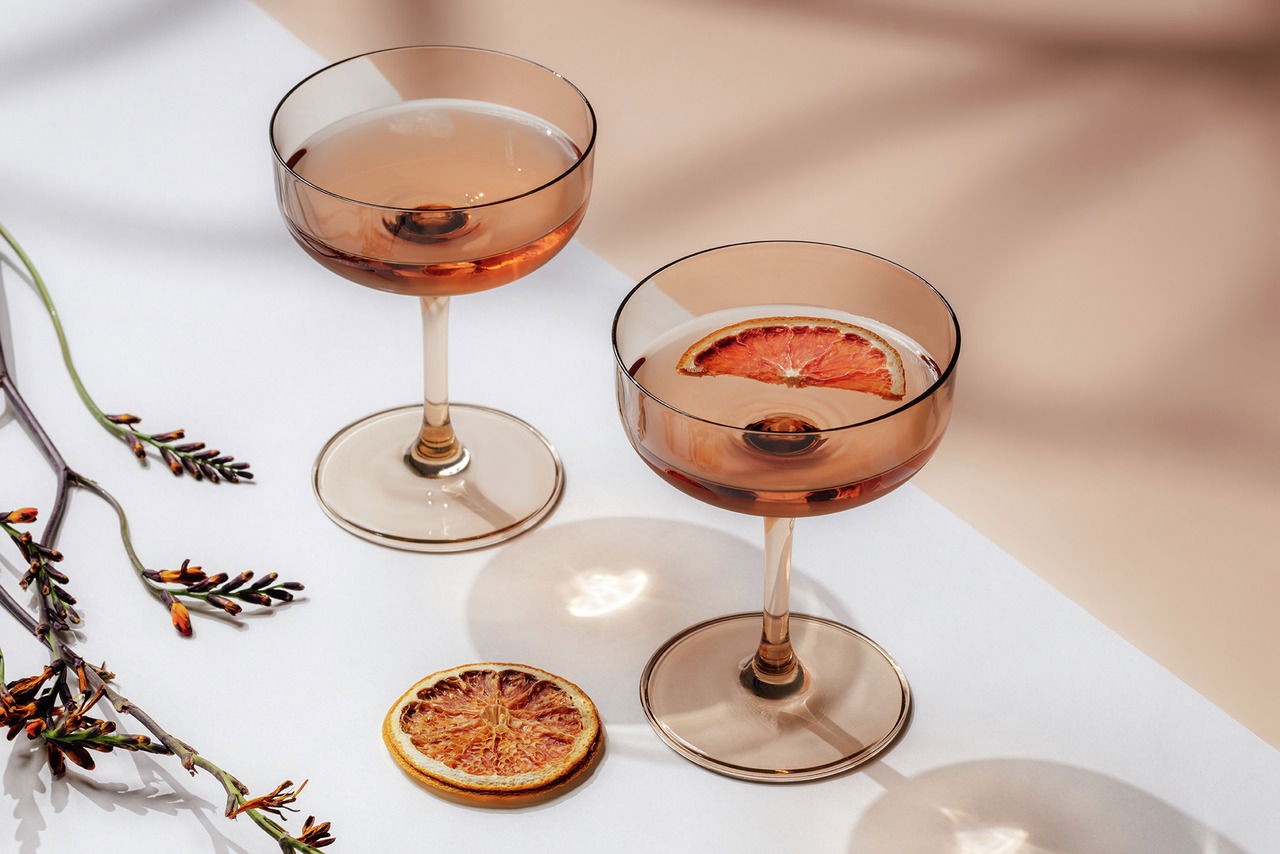 Zwei elegante Sekt- und Dessertschalen aus bernsteinfarbenem Glas, gefüllt mit Getränken und einer getrockneten Orangenscheibe, auf einer weißen Oberfläche mit dekorativen Zweigen, aus der Vogelperspektive fotografiert.