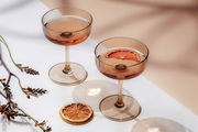 Zwei elegante Sekt- und Dessertschalen aus bernsteinfarbenem Glas, gefüllt mit Getränken und einer getrockneten Orangenscheibe, auf einer weißen Oberfläche mit dekorativen Zweigen, aus der Vogelperspektive fotografiert.