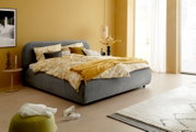 Graues Polsterbettgestell in einem modern eingerichteten Schlafzimmer mit gelber Bettwäsche und Dekoration, seitliche Perspektive.