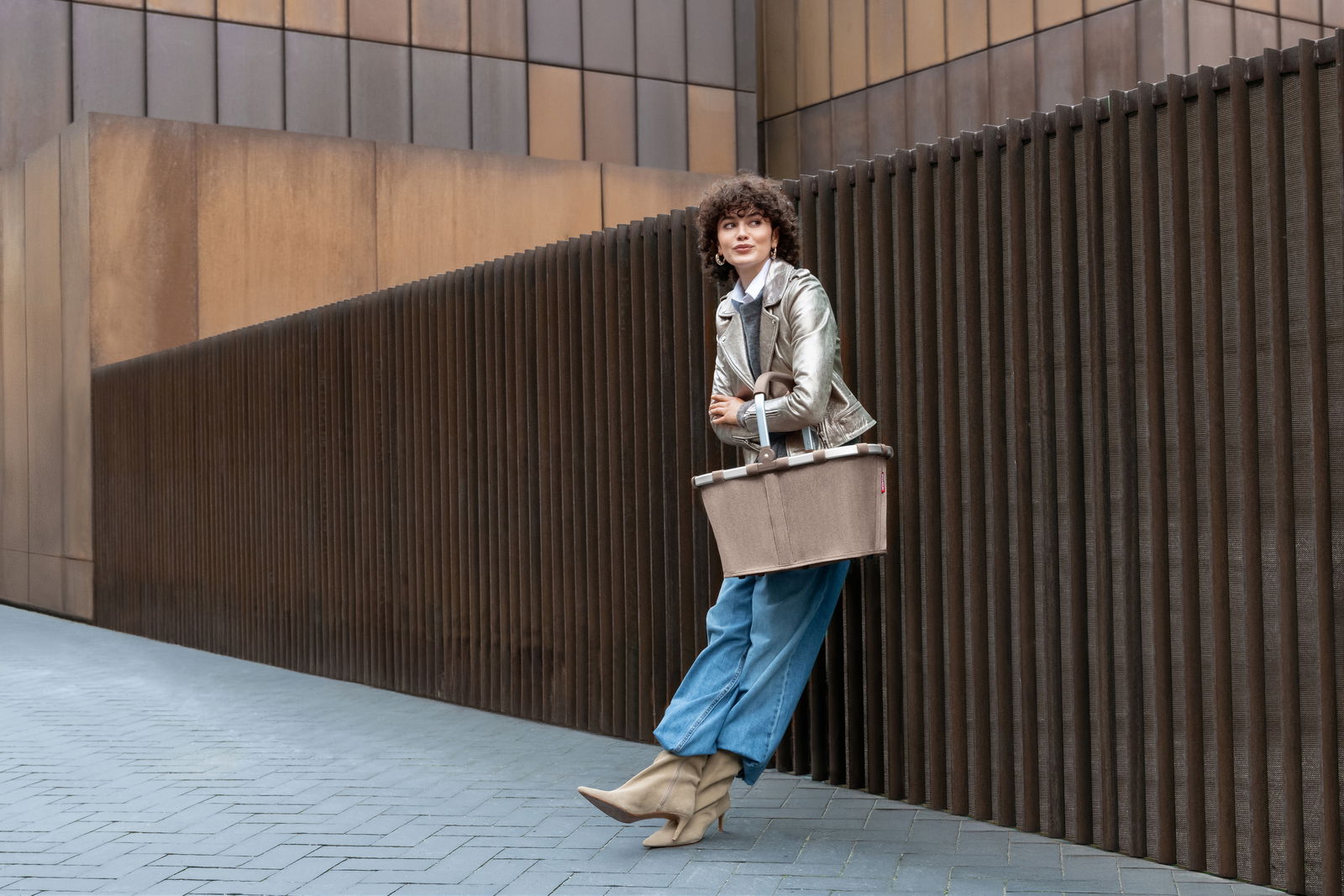 Frau lehnt an einer modernen Wand und hält einen braunen Einkaufskorb in der Hand, seitliche Perspektive.