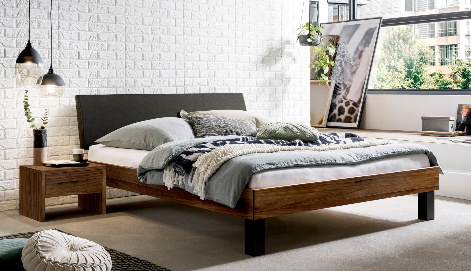 Modern houten tweepersoonsbed met een donkergrijs gestoffeerd hoofdeinde en een zwart voeteneinde, opgemaakt met grijs beddengoed in een lichte slaapkamer – perspectief: 3/4-vooraanzicht vanaf links.