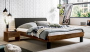 Modern houten tweepersoonsbed met een donkergrijs gestoffeerd hoofdeinde en een zwart voeteneinde, opgemaakt met grijs beddengoed in een lichte slaapkamer – perspectief: 3/4-vooraanzicht vanaf links.