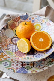 Frische Orange, halbe Orange und halbe Zitrone in einer bunten Schale mit Patchwork-Muster auf Holztisch, fotografiert in leicht schräger Draufsicht
