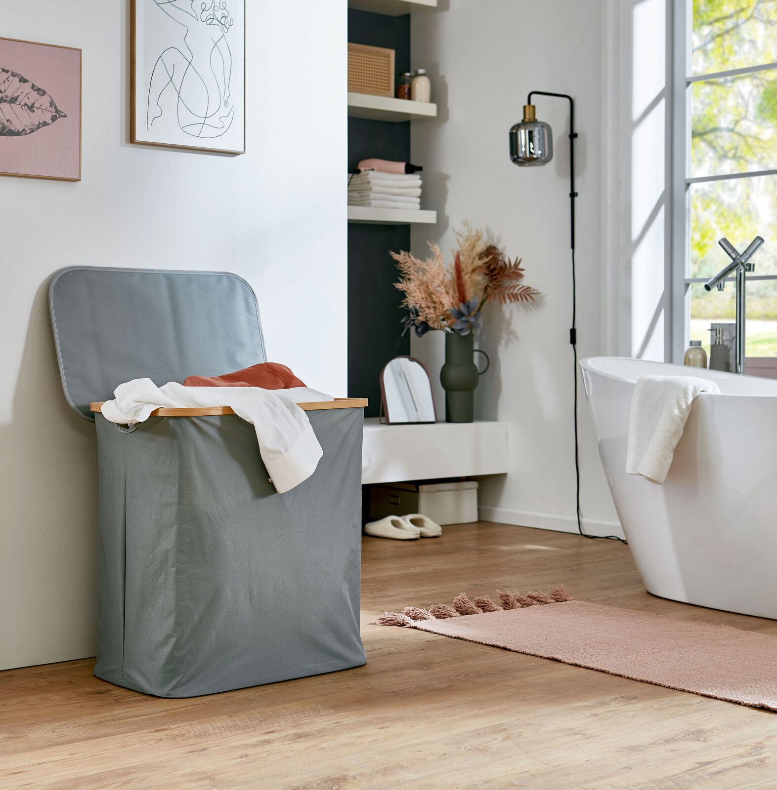 Grauer Wäschekorb mit Deckel und Holzrahmen in einem hellen Badezimmer neben einer freistehenden Badewanne, seitliche Perspektive.