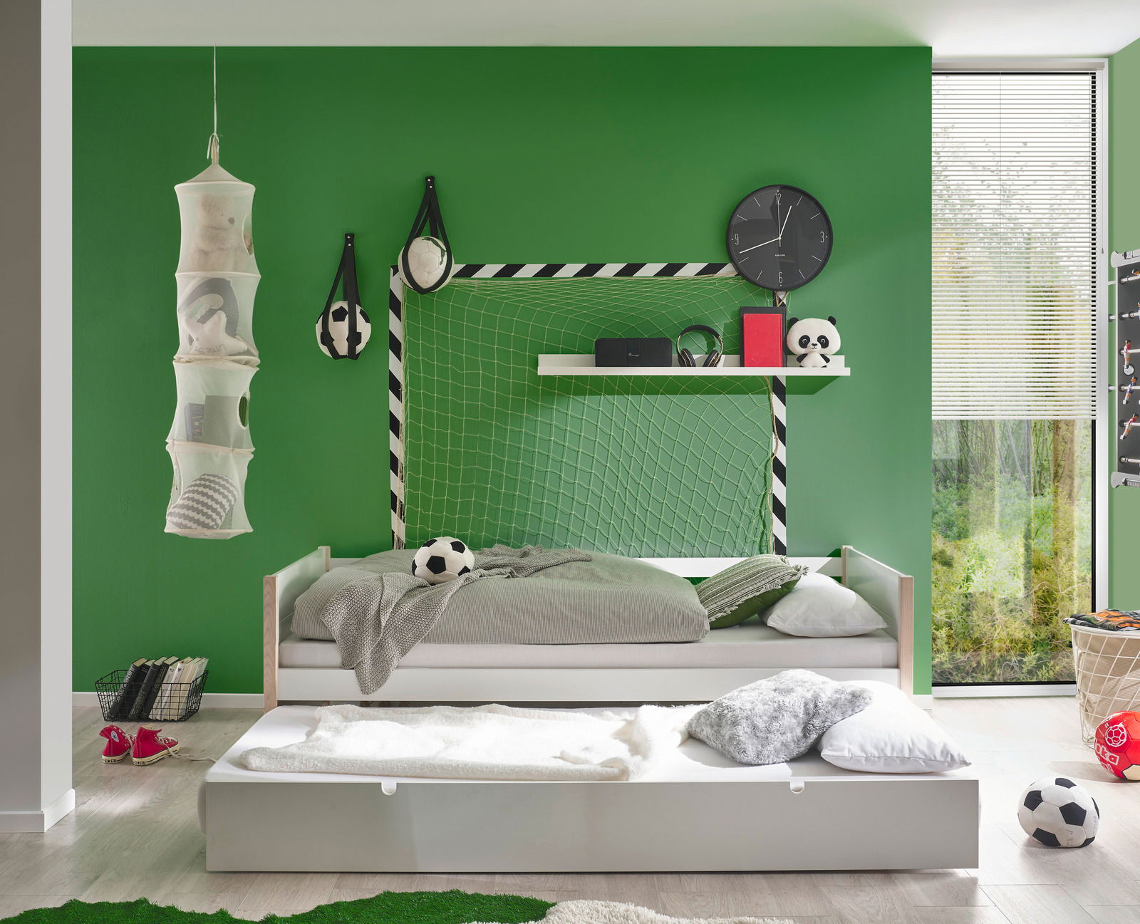 Jugendbett in einem Kinderzimmer mit grün gestrichener Wand, Fußball-Dekoration und einem ausziehbaren Bettkasten, seitliche Perspektive.