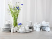 Weißes Porzellan-Geschirrset mit Tellern, Schalen, Tassen, Kanne und Schüssel auf hellem Holztisch, dekoriert mit Glasvase und blauen Blumen, frontal aus leicht erhöhter Perspektive vor weißem Hintergrund