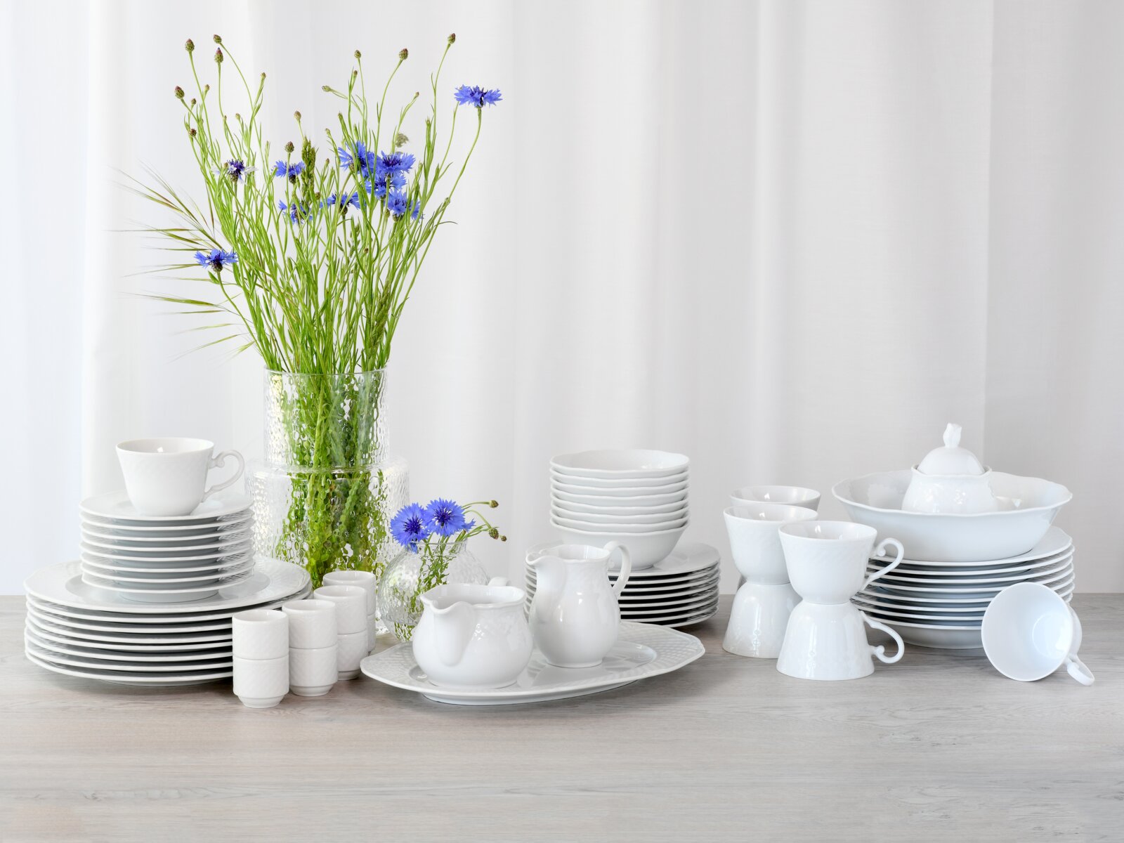 Weißes Porzellan-Geschirrset mit Tellern, Schalen, Tassen, Kanne und Schüssel auf hellem Holztisch, dekoriert mit Glasvase und blauen Blumen, frontal aus leicht erhöhter Perspektive vor weißem Hintergrund