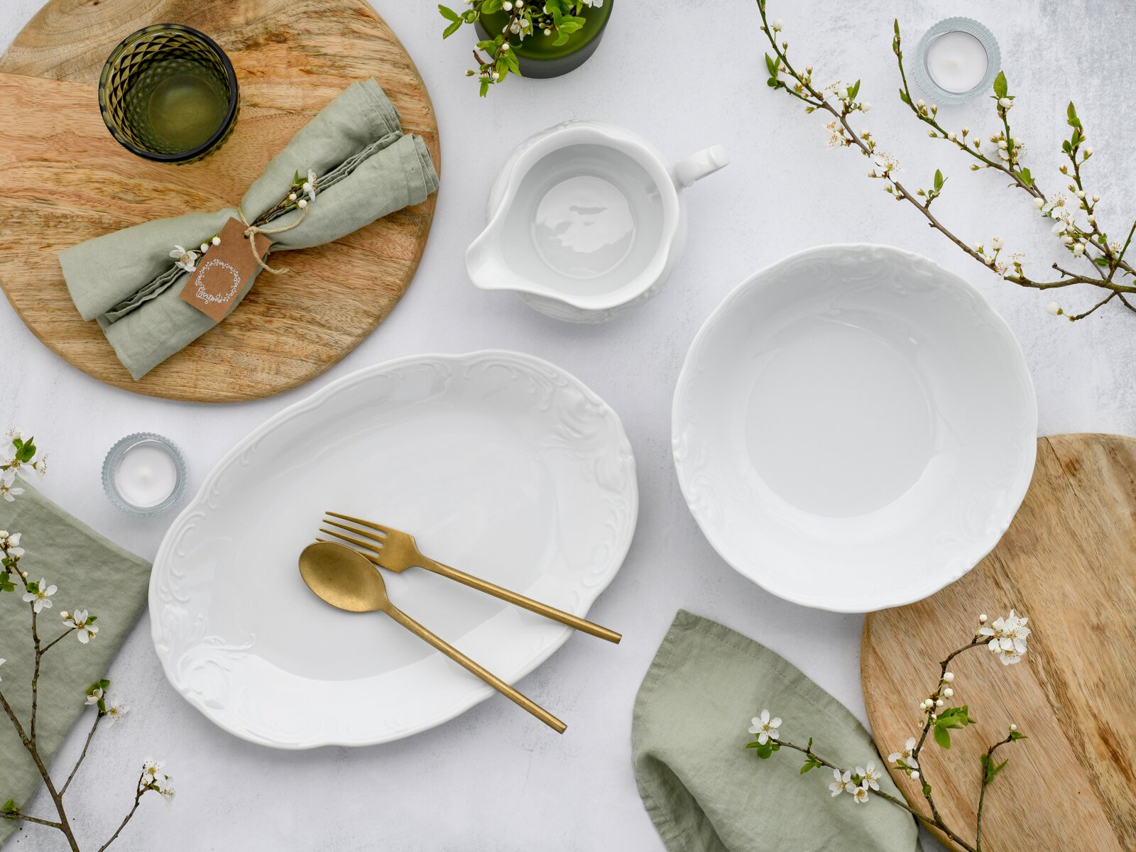 Draufsicht auf ein elegantes weißes Porzellan-Servierset mit ovaler Platte, Schale und kleiner Sauciere mit Reliefdekor, dekoriert mit goldfarbenem Besteck, Leinenservietten, Holzbrett, Kerzen und Frühlingszweigen auf hellem Hintergrund.