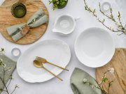 Draufsicht auf ein elegantes weißes Porzellan-Servierset mit ovaler Platte, Schale und kleiner Sauciere mit Reliefdekor, dekoriert mit goldfarbenem Besteck, Leinenservietten, Holzbrett, Kerzen und Frühlingszweigen auf hellem Hintergrund.