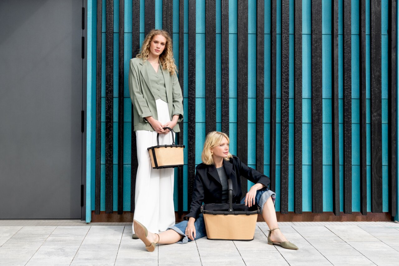 Zwei Frauen mit stilvollen, modernen Tragetaschen vor einer blau-schwarzen Wand. Eine Frau steht und hält eine kleinere Tasche, während die andere sitzt und eine größere Tasche neben sich hat. Frontale Perspektive.